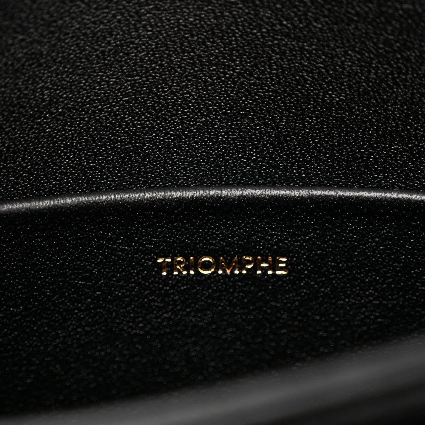 Black leather bag surface with 'TRIOMPHE' embossed lettering and fine grain texture.